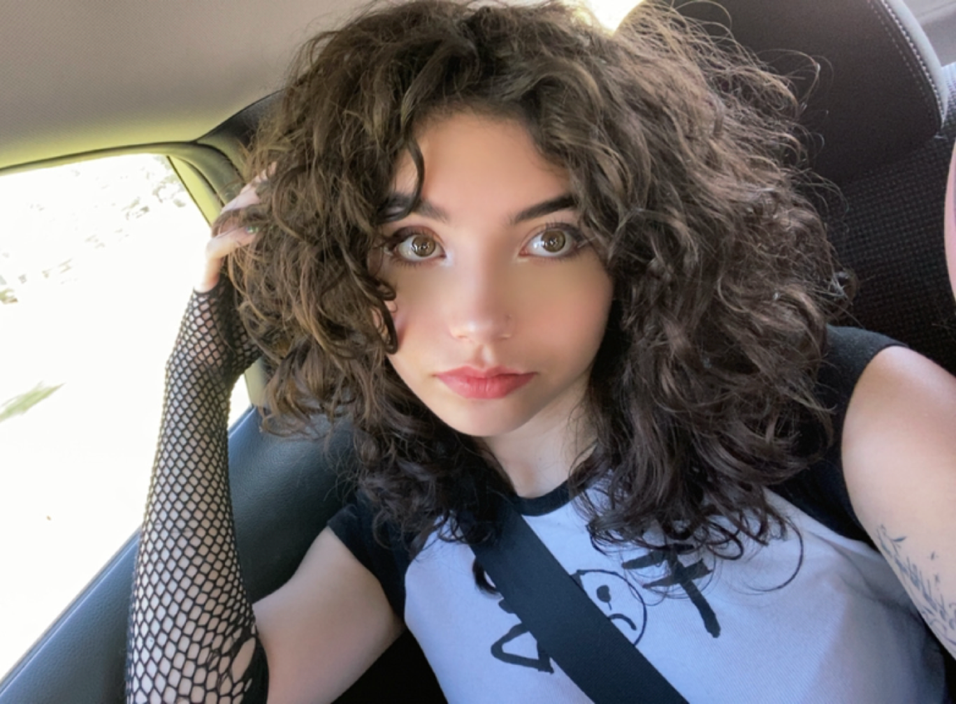 A selfie of Hailee wearing a white T-shirt.She is looking at the camera with a serious expresion, and her hand is placed upon her head holding her brown curly hair.